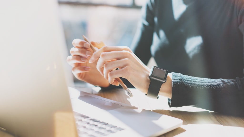 A woman's hands rest on a desk in front of a laptop, holding a pencil