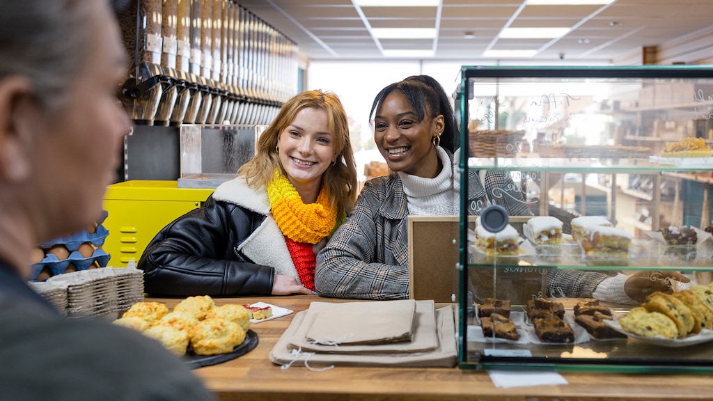 Over the shoulder shot of a cafe worker helping two friends browsing a cafe that promotes sustainable living in the North East of England. The store serves delicious sweet vegan treats, the two young female friends are viewing the treats on display.