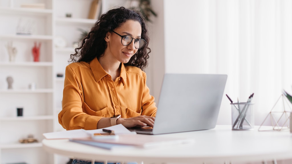 Middle Eastern Lady Using Laptop Working Online Wearing Eyewear Sitting At Workplace In Modern Office. Remote Job, Technology And Career Profession Concept. Side View