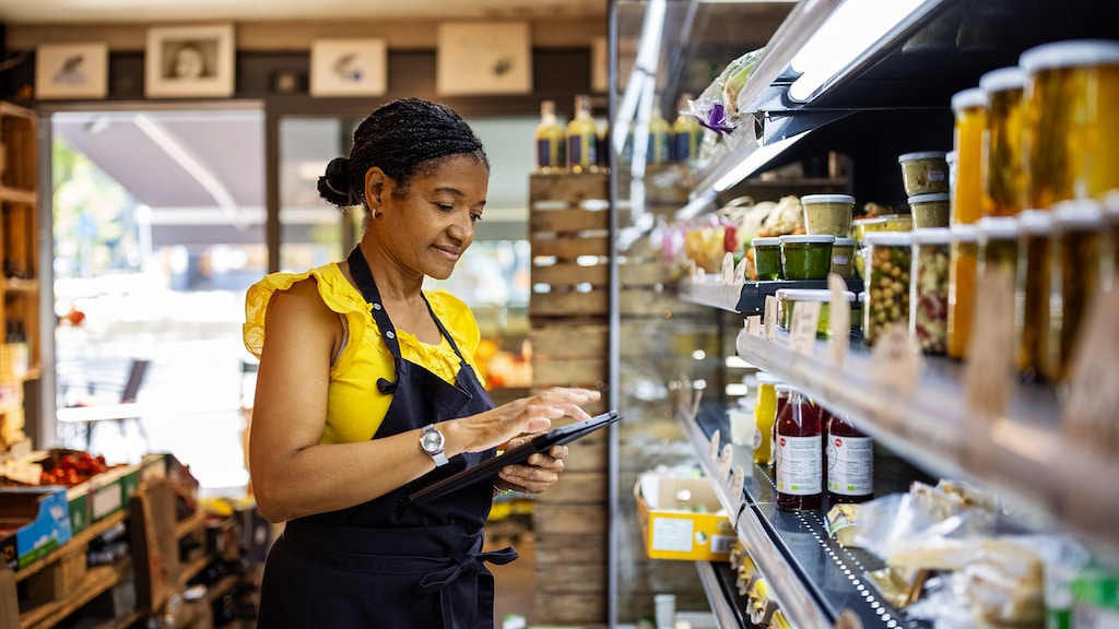 Confident saleswoman checking inventory in grocery store. Female owner is using digital tablet in store. She is working in delicatessen.